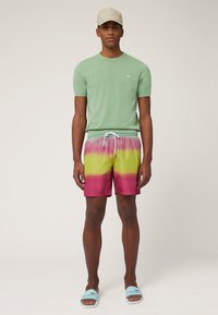 Groene korte mouwen t-shirt, zwemshorts met een roze-geel verloop en trekkoord, en blauwe slippertjes. Casual zomeroutfit met logo-details.