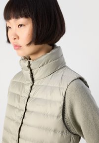 Ung kvinne med kort mørkt hår, iført en lys beige quiltet vest over en strikket genser, som ser litt nedover mot en ensfarget bakgrunn.
