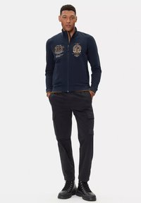 Felpa navy con zip e dettagli ricamati sul davanti, abbinata a pantaloni cargo neri e stivali neri con lacci. Tessuto liscio, design casual.