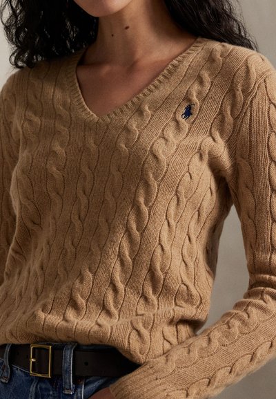 Žena nosí béžový pletený sveter s výstrihom do V a modro vyšívaným logom, v kombinácii s modrými džínsami a čiernym opaskom so zlatou sponou.