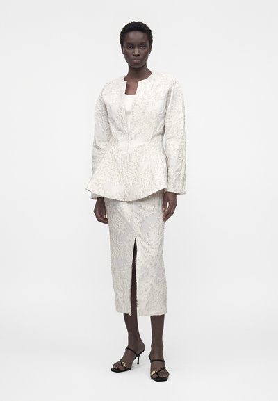 Γυναίκα που φοράει υφασμένο κρεμ σακάκι με μέση πεπλούμ και ταιριαστό μίντι φούστα με εμπρόσθια σχισμή, συνδυασμένο με μαύρα σανδάλια με τακούνι.