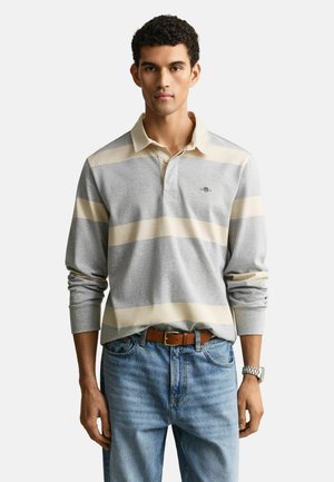 Man draagt een grijs-beige gestreept poloshirt met lange mouwen, blauwe spijkerbroek, een bruine riem en een zilveren horloge, staand tegen een witte achtergrond.