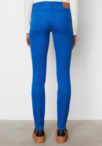 Jeans skinny azul con una textura suave, dos bolsillos traseros y un parche de cuero marrón en la cintura, combinados con zapatos de plataforma negros.