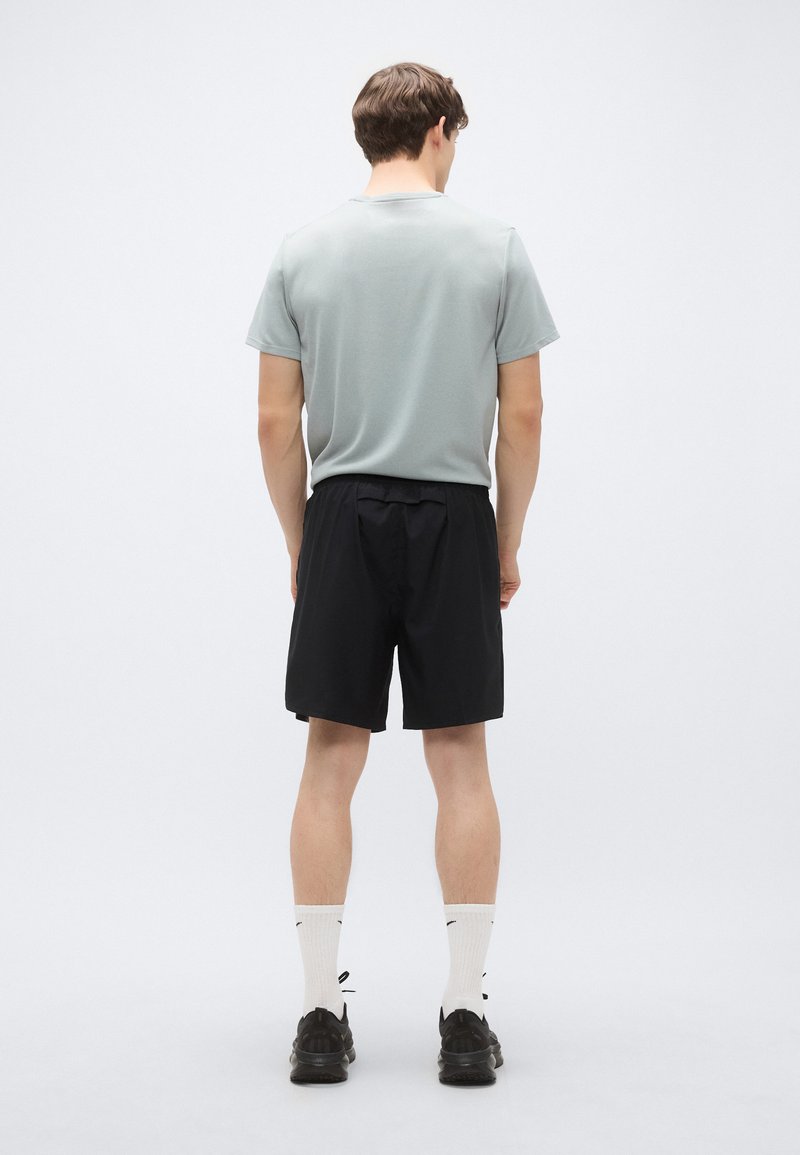 Graues kurzärmliges Hemd kombiniert mit schwarzen Shorts. Das Modell trägt weiße Socken mit schwarzen Akzenten und schwarze Sportschuhe. Einfaches Design, lässige Passform.