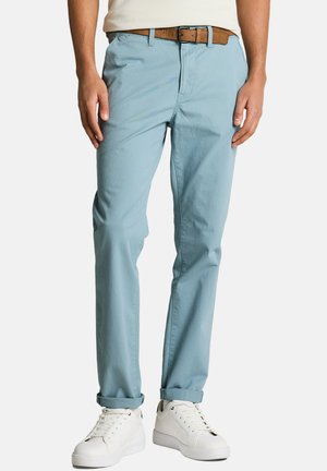 Homme portant un pantalon bleu clair retroussé avec une ceinture marron et des baskets blanches, debout devant un fond uni.