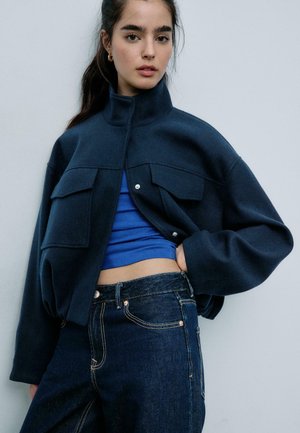 Mlada ženska s temnimi lasmi, oblečena v kratko mornarsko modro jakno, modro kratko majico in temne kavbojke, ki pozira na enobarvnem svetlem ozadju.