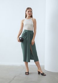 Cream tank top paired with a green wrap skirt featuring a front slit, held with a knot detail. Black handbag in hand, wearing black heeled sandals.