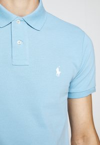 Ljust blå polo med två vita knappar och en liten vit broderad logotyp av en polo-spelare på en häst på bröstet.