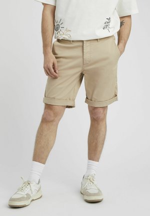 Shorts beiges en tissu léger, avec des revers roulés, des poches latérales et une coupe régulière, associés à des baskets blanches et des chaussettes rayées.