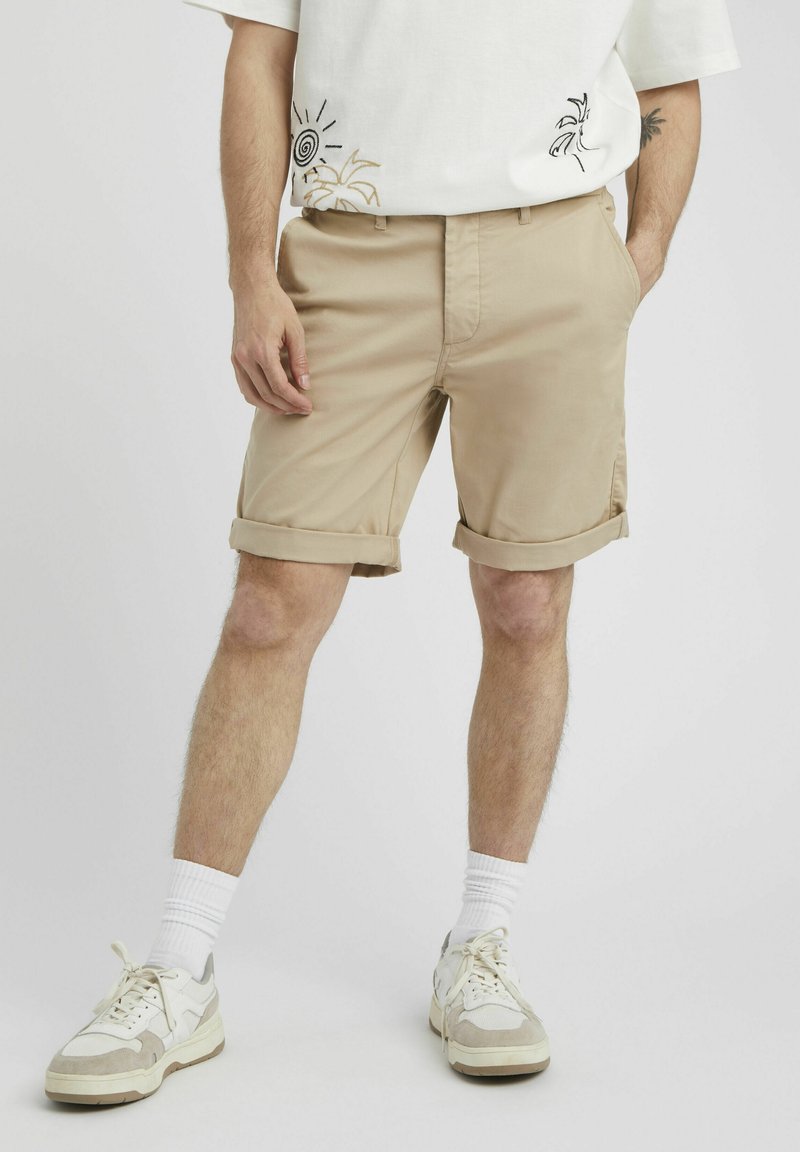 Shorts beiges en tissu léger, avec des revers roulés, des poches latérales et une coupe régulière, associés à des baskets blanches et des chaussettes rayées.