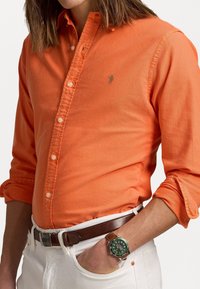 Orange skjorta med knappar och en diskret logotyp, bärs tillsammans med vita byxor och ett brunt läderbälte. En grön klocka syns på handleden.