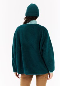 Osoba s teplou fleecovou bundou v teal farbe, teal pletenou čiapkou, svetlom modrým rolákom a hnedými nohavicami, zobrazená zozadu na jednoduchom pozadí.