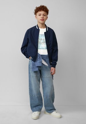 Veste de pilote navy sur un t-shirt graphique blanc, associée à un jean large délavé bleu clair et des baskets blanches. Look décontracté en couches.