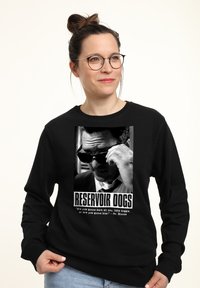 Eine Frau, die einen schwarzen Sweatshirt trägt, hat ein auffälliges Grafikdesign eines Mannes mit Sonnenbrille, zusammen mit dem Text "RESERVOIR DOGS" und einem Zitat darunter.