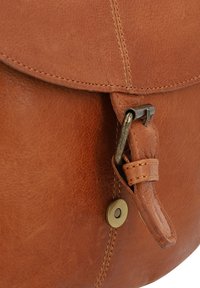 Ruda odinė rankinė su apvalia forma, turinti sulankstomą dangtelį, žalvario furnitūrą ir matomus siuvimo detalių. Tekstūruotas paviršius.