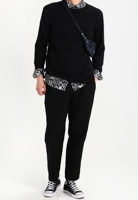 Persona che indossa un maglione nero a pois sopra una camicia a fantasia, pantaloni neri, sneakers alte nere e una piccola borsa a tracolla.