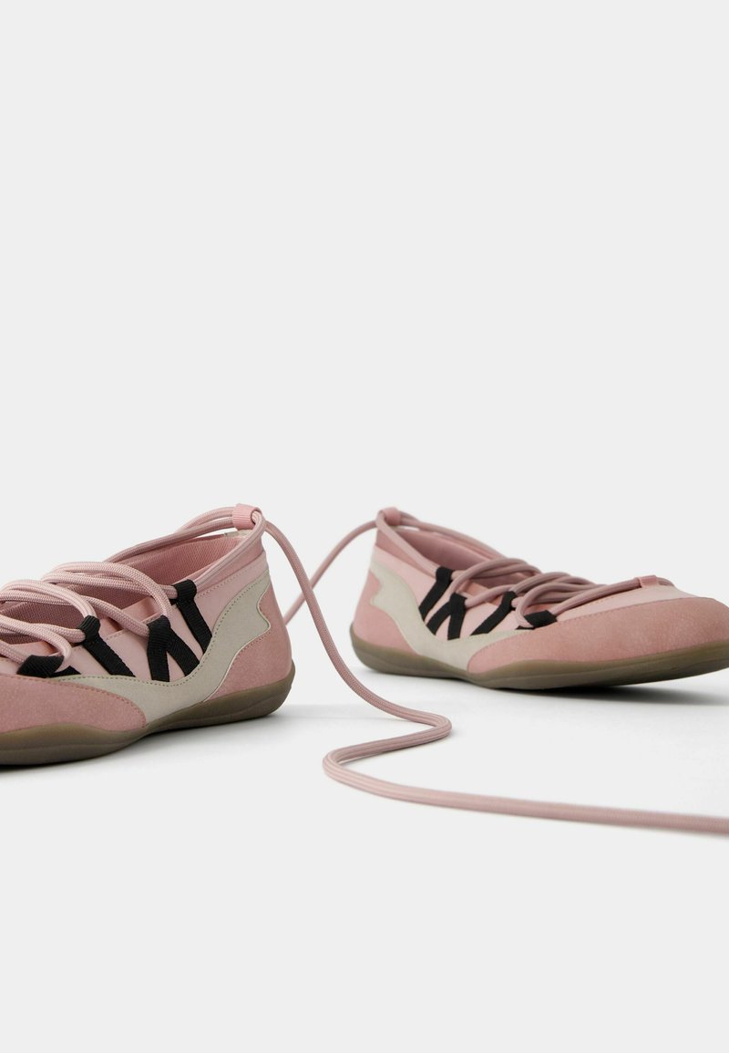Zapatillas en tonos rosa y beige con una textura suave, que presentan cordones negros y una suela flexible. Los cordones están atados de manera suelta, con uno que se extiende por el suelo.
