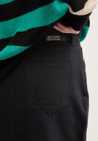 Aukšto liemens juodos džinsinės kelnės su galine kišene ir etikete, pažymėta "DENIM TOM TAILOR." Medžiaga atrodo lygi ir patvari.