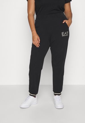 Osoba nosząca czarne spodnie dresowe z logo EA7 Emporio Armani na udzie i białe tenisówki stojąca na białym tle.