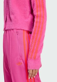 Fuzzy pink sweater with orange stripes on sleeves, paired with pink pants featuring a zip pocket and logo. Both have a soft texture.