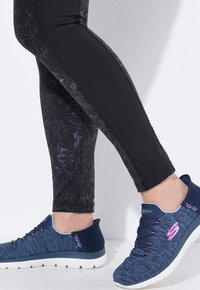 Blå sportskor med texturerat tyg, rosa detaljer och vita sulor, tillsammans med svarta leggings med ett blommönster och utskriftsdesign.