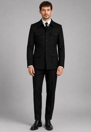Homme portant une veste noire boutonnée avec quatre poches à rabat, un pantalon noir, une chemise blanche, une cravate noire et des chaussures habillées noires, debout devant un fond gris.