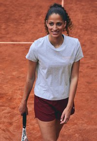 Grå kortärmad t-shirt med en rosa logotyp, i kombination med vinröda shorts, stående på en tennisbana av röd lera, hållande ett tennisslag.