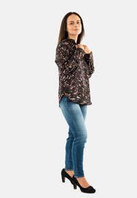 Mujer de pie con una camisa de manga larga con estampado, jeans azules y tacones negros ajustándose el cuello contra un fondo claro y liso.