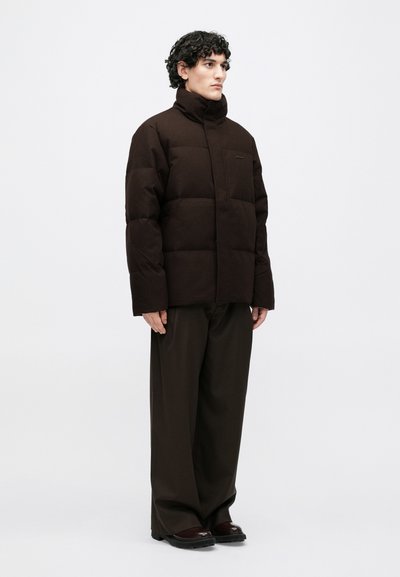 Brūna pūkaina jaka ar augstu apkakli, kurā ir taisnstūrveida šūtas daļas. Modelis valkā arī tumšas, platas bikses un tumšus apavus.