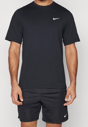 Mann trägt schwarzes Nike-T-Shirt mit kurzen Ärmeln und passende schwarze Nike-Shorts, steht vor einem schlichten hellen Hintergrund.