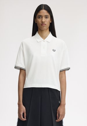 Femme aux longs cheveux foncés portant un polo blanc court avec des bordures noires et une jupe plissée noire sur un fond uni.