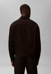 Hombre con chaqueta vaquera negra y vaqueros negros de pie frente a un fondo liso de color gris claro, mostrando la parte trasera de la chaqueta.