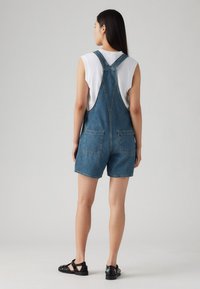 Denim kratke naramnice v srednje modri barvi, z nastavljivimi naramnicami in dvema zadnjima žepoma. Obuti čez belo majico brez rokavov in črne sandale.