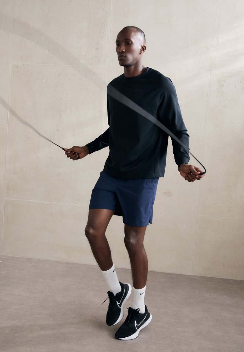 Camisa negra de manga larga, pantalones cortos azul marino y zapatillas negras con detalles en blanco. Cuerda de saltar en la mano. Fondo minimalista, texturas suaves.