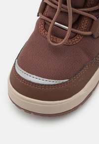 Zapatilla de tela marrón con parte superior texturizada, acento plateado y suela de goma beige. Presenta cordones marrones y un detalle del logo en el lateral.