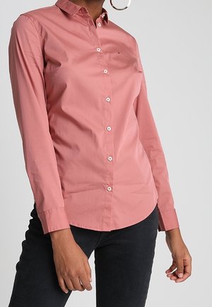 Woman wearing a buttoned-up long-sleeve pink shirt and black pants, with large hoop earrings visible on one side.