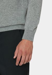 Grauer Strickpullover mit geripptem Bündchen, gezeigt neben einer Hand, die auf dunklen Hosen ruht. Weiche Textur, schmaler Schnitt und minimalistischer Stil.