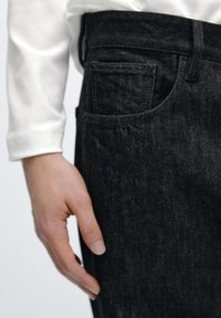 Mørk denimjeans med en tætsiddende pasform, der har en baglomme, diskrete syninger og en glat tekstur. En hvid langærmet skjorte kan ses.