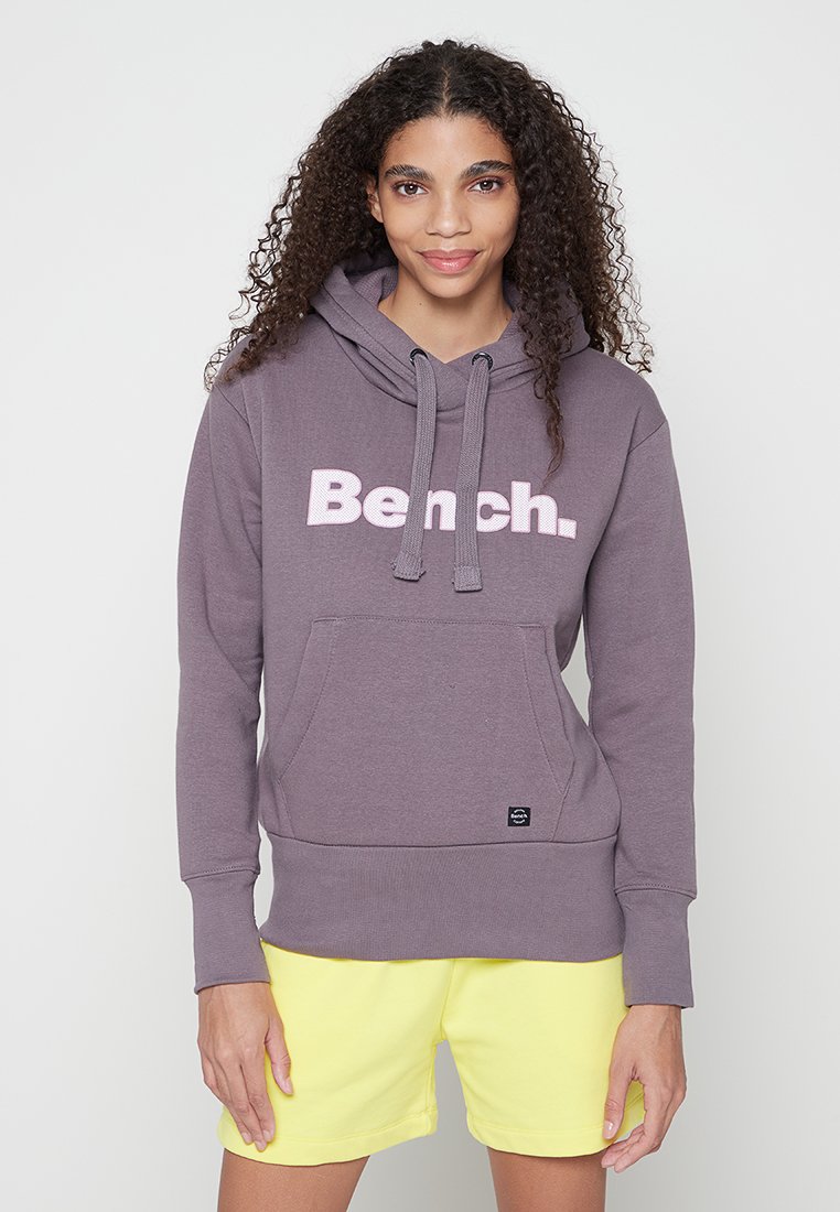 Lila Kapuzensweatshirt mit weicher Textur, ausgestattet mit einem weißen "Bench."-Logo. Enthält eine Vordertasche und Kordeln, kombiniert mit gelben Shorts.