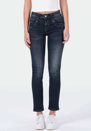 Vrouw draagt donker gewassen skinny jeans, witte sneakers en een wit cropped topje, staand met ontspannen armen langs haar zij.