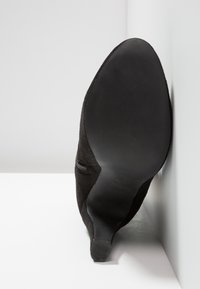 Chaussure à talon haut en daim noir avec un bout pointu et une fermeture éclair latérale, dotée d'une semelle en caoutchouc texturée et d'une silhouette élégante.