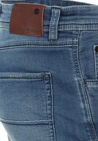 Denim-Jeans in einer mittelblauen Waschung, mit einem braunen Lederpatch, das "CASA MODA" bezeichnet, mit sichtbaren Nähten und mehreren Taschen.