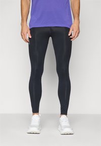 Un homme portant des leggings de sport noirs, des chaussures de course blanches, et un t-shirt violet, se tenant devant un fond clair et uni.