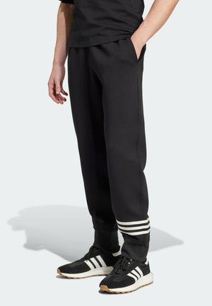 Personne portant un jogging noir avec des bandes blanches près des poignets, associé à des baskets Adidas noires et blanches, debout devant un fond uni.