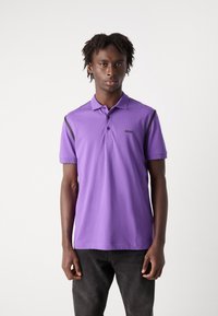 Μωβ πόλο μπλουζάκι από μαλακό ύφασμα, με τριπλό κουμπί, κοντά μανίκια και μαύρες λεπτομέρειες στους ώμους.