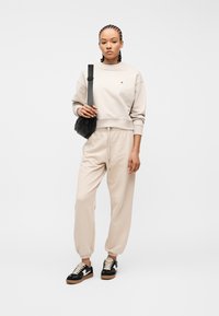 Νεαρή γυναίκα με πλεξούδες, φοράει μπεζ φούτερ και φόρμες, μαύρη τσάντα χιαστί και μαύρα και λευκά αθλητικά παπούτσια, στέκεται όρθια.