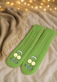 Grüne Socken mit Zopfmuster-Struktur und einem Cartoon-Gesicht mit großen Augen und sichtbaren Zähnen, auf einem weichen, neutralen Hintergrund platziert.