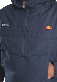 Felpa con cappuccio blu navy con zip frontale e grande tasca anteriore. Presenta il logo "ellesse" e un emblema del sole ricamato a blocchi di colore.