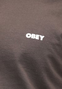 Brun t-shirt med korte ærmer, der har et hvidt "OBEY" logo på brystet. Lavet af blødt bomuldsstof med en glat tekstur.