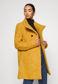 Cappotto di un giallo brillante con una texture, dotato di un ampio colletto e caratterizzato da due grandi bottoni, indossato sopra un dolcevita nero e jeans blu.